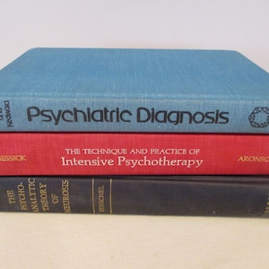 Op de afbeelding: Drie op elkaar gestapelde hardcover boeken. Het bovenste boek is blauwgroen en heeft de titel "Psychiatric Diagnosis". Het middelste boek is rood en leest "The Technique and Practice of Intensive Psychotherapy". Het onderste boek is donkerblauw.