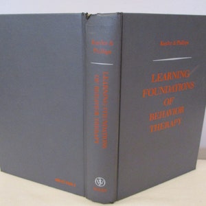 Op de afbeelding: Een grijs hardback boek getiteld "Learning Foundations of Behavior Therapy" door Kazdin & Phillips. Het boek is uitgegeven door Wiley.