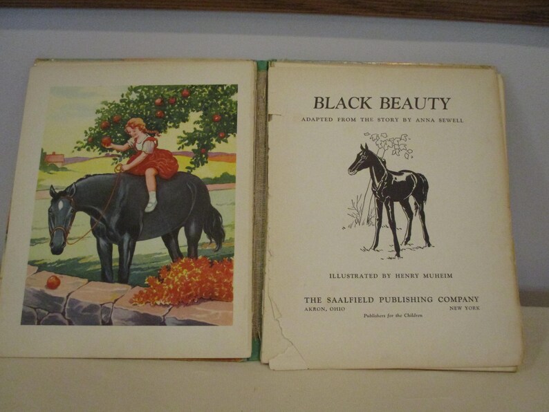May include: An open vintage book titled "Black Beauty" with illustrations. The left page shows a girl in a red dress on a black horse under an apple tree. The right page has a black horse illustration and the book's title.