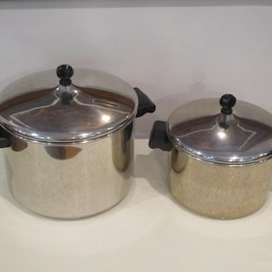 May include: Two stainless steel pots with matching lids, each featuring black plastic side handles and a black knob on the lid. The pots have a polished, reflective finish and a cylindrical shape, with one pot noticeably larger than the other.
