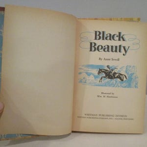May include: An open book titled "Black Beauty" by Anna Sewell. The cover features a blue and gold floral pattern. The book's title page shows a horse and rider illustration in blue and white.