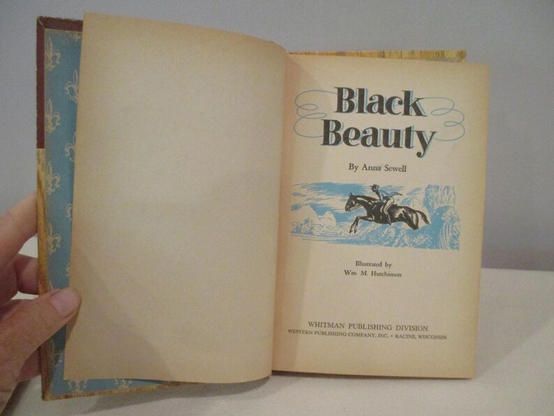 May include: An open book titled "Black Beauty" by Anna Sewell. The cover features a blue and gold floral pattern. The book's title page shows a horse and rider illustration in blue and white.