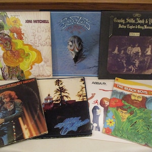Può includere: Una collezione di dischi in vinile vintage con artisti come Joni Mitchell, The Eagles, Crosby, Stills, Nash & Young, ABBA e The Beach Boys. Le copertine dei dischi presentano una varietà di colori e design, tra cui un motivo floreale psichedelico, un paesaggio blu e bianco, una fotografia in bianco e nero e un'illustrazione colorata di una scena di spiaggia.