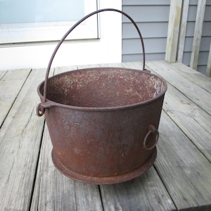 Może przedstawiać: Rustykalny kocioł żeliwny z metalową rączką i pierścieniem po boku. Kocioł stoi na drewnianym tarasie.