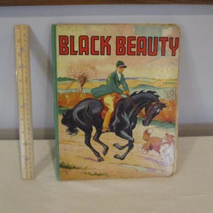 May include: Vintage hardcover book titled "BLACK BEAUTY." The cover features an illustration of a rider on a black horse, with a dog running alongside. The book has a green border and a wooden ruler is next to the book.