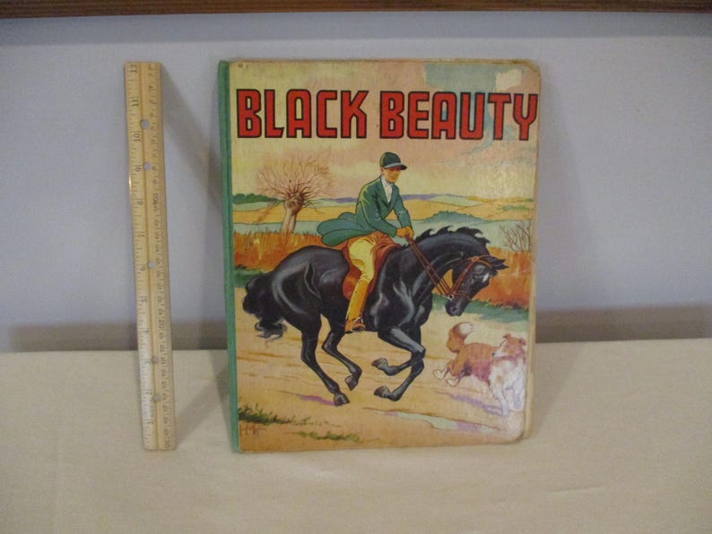 May include: Vintage hardcover book titled "BLACK BEAUTY." The cover features an illustration of a rider on a black horse, with a dog running alongside. The book has a green border and a wooden ruler is next to the book.