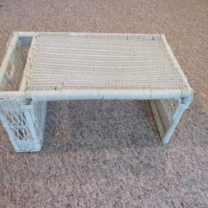 Puede incluir: Una bandeja de cama de mimbre blanco con una parte superior rectangular y patas en ángulo. La bandeja tiene un diseño tejido y un pequeño compartimento lateral. La bandeja está diseñada para ser utilizada en una cama u otra superficie.