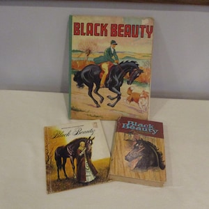 May include: Three vintage copies of the book "Black Beauty" are displayed. The largest book features a rider on a black horse with dogs. The other two books have illustrations of a black horse and the title "Black Beauty".