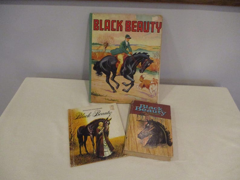 May include: Three vintage copies of the book "Black Beauty" are displayed. The largest book features a rider on a black horse with dogs. The other two books have illustrations of a black horse and the title "Black Beauty".
