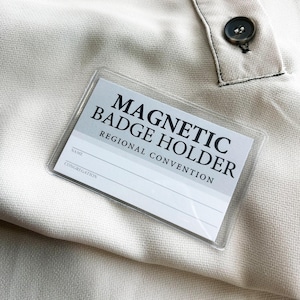 May include: A clear rectangular magnetic badge holder with a white insert featuring the black text MAGNETIC BADGE HOLDER, REGIONAL CONVENTION, NAME, and CONGREGATION. The badge holder is positioned on a light beige fabric with a dark button visible.