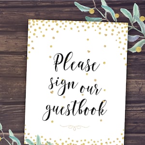 May include: A white sign with gold confetti and the text "Please sign our guestbook" in elegant black script. The sign is decorated with gold dots and a decorative flourish. The background is a dark wood texture.