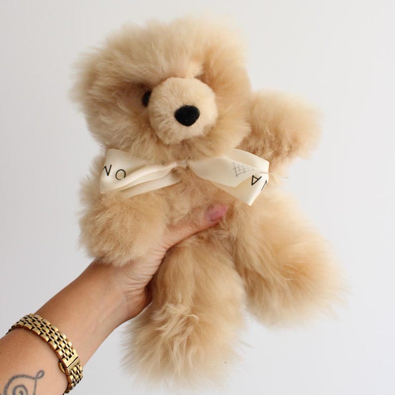 Pu&ograve; includere: Un orsacchiotto di peluche beige chiaro dall'aspetto soffice e testurizzato. L'orso presenta naso e occhi marrone scuro ed &egrave; decorato con un nastro color crema annodato al collo, che mostra una scritta parziale in lettere nere.