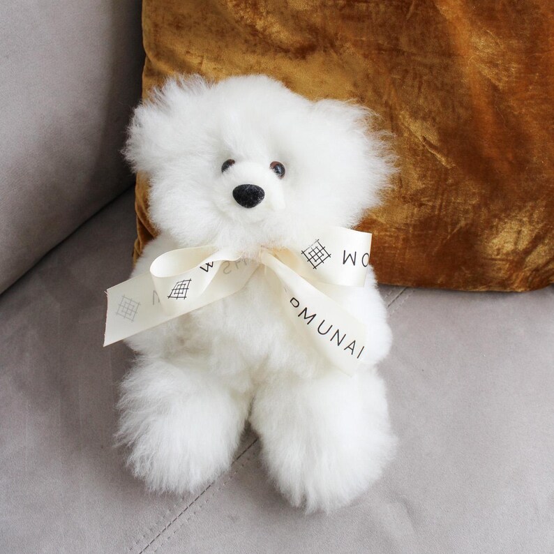 Pu&ograve; includere: Un soffice orsacchiotto bianco con naso nero e occhi scuri. L'orso presenta un nastro color crema legato attorno al collo, stampato con il testo MUNAI e un piccolo motivo a griglia. Il peluche ha una consistenza morbida e arruffata su tutto il corpo.