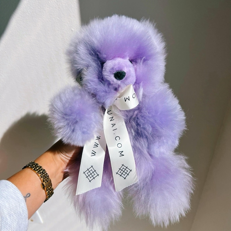 Pu&ograve; includere: Un soffice orsacchiotto color lavanda dall'aspetto morbido e testurizzato. Un nastro bianco &egrave; legato attorno al collo, con il testo stampato 'WWW.UNAI.COM' insieme a un piccolo logo geometrico. L'orso ha un naso scuro e orecchie arrotondate.