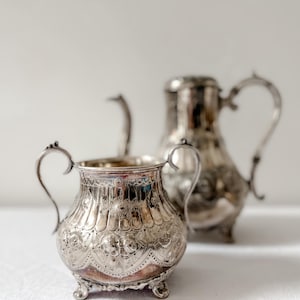 Puede incluir: Un azucarero de plata con detalles ornamentados y dos asas, junto con una tetera de plata con un diseño similar.
