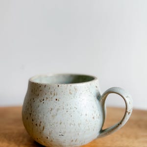 May include: A speckled, pale blue ceramic mug with a rounded body and a curved handle. The mug has a slightly textured surface and a light brown wooden surface in the background. The mug is designed for holding hot beverages.