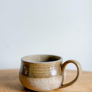 Puede incluir: Una taza de cerámica hecha a mano con un cuerpo redondeado y un asa robusta. La taza presenta un diseño de dos tonos, con una base beige claro y una sección superior marrón más oscura. El borde tiene una fina banda clara. La taza está sobre una superficie de madera.