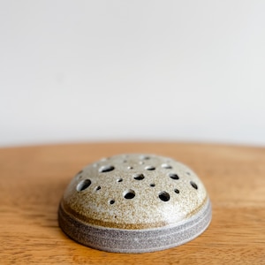 May include: A ceramic flower frog with a speckled, light brown and grey finish. The frog has a rounded shape with numerous holes for arranging flower stems. The base is a darker grey colour, and it sits on a wooden surface.