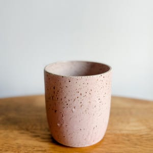 May include: A pink ceramic cup with a speckled texture and a rounded base. The cup has a smooth rim and is covered in small, dark brown spots. The cup is on a wooden surface.