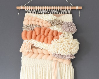 May include: A woven wall hanging with a cream, peach, and light brown colour scheme. The hanging features a variety of textures, including pom-poms, tassels, and braids. The hanging is attached to a wooden dowel.