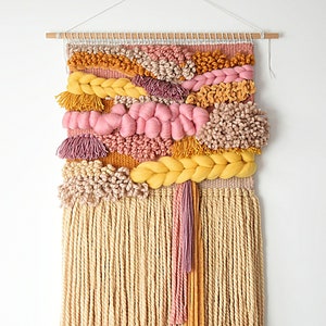 Könnte beinhalten: Ein gewebter Wandbehang mit einem farbenfrohen abstrakten Design. Der Behang besteht aus rosa, gelbem und beigefarbenem Garn in verschiedenen Texturen, darunter Pompons, Zöpfe und Fransen.
