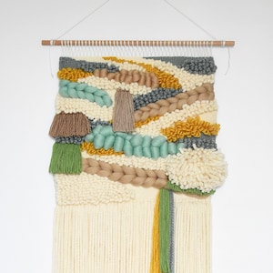 Op de afbeelding: Een geweven wandkleed met verschillende texturen en kleuren, waaronder beige, bruin, groen, grijs en geel. Het kleed heeft een franje rand en is gemaakt van garen.