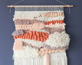 May include: A woven wall hanging featuring a mix of textures and colours, including peach, coral, and cream. The hanging has a fringe border and is crafted from natural fibres.
