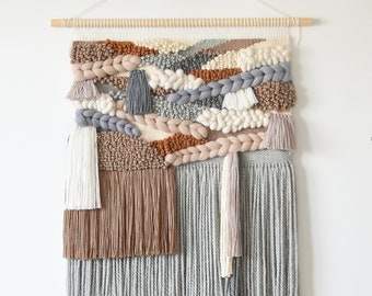 May include: A woven wall hanging with a variety of textures and colors, including brown, beige, gray, and white. The hanging features a fringe border and is made with yarn.