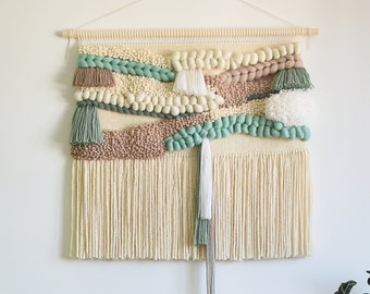 May include: A woven wall hanging with a beige background and green, brown, and white accents. The hanging features a variety of textures, including pom-poms, fringe, and woven patterns.