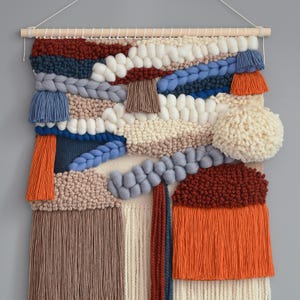 May include: A woven wall hanging with a variety of textures and colors. The piece features a wooden dowel at the top for hanging, with sections of white, beige, blue, and orange yarn. Braided and fringed elements add dimension.
