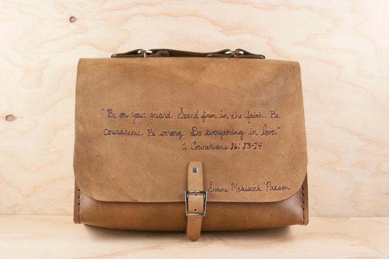 May include: A brown leather satchel with a buckle closure and a quote from 1 Corinthians 16:13-14 inscribed on the front. The quote reads: "Be on your guard. Stand firm in the faith. Be courageous. Be strong. Do everything in love." The bag is signed "Shane McKissick Parson".