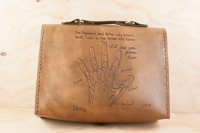 May include: A brown leather bag with a handprint design and the text "The husband and father who knows best, looks to the Father who knows ALL. I'll love you forever! Kara Sienna Isaiah Me Kaylyn 2019 Elana"