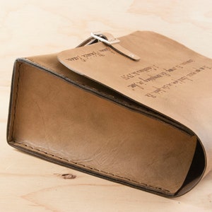 May include: A brown leather satchel bag with a flap closure and a metal buckle. The bag has a strap and is engraved with text.