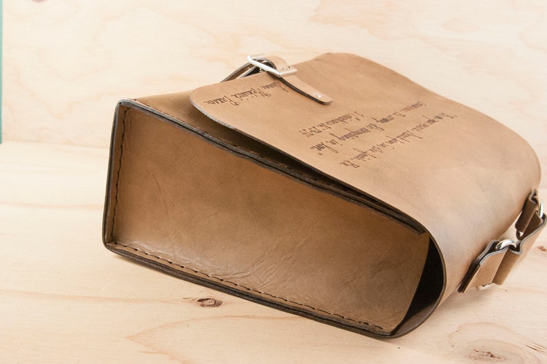 May include: A brown leather satchel bag with a flap closure and a metal buckle. The bag has a strap and is engraved with text.