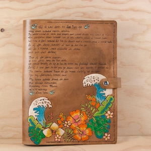 Puede incluir: Un diario de cuero marrón con un mensaje escrito a mano y un diseño pintado de olas y flores tropicales. El mensaje dice: "Si alguna vez tuvieras que irte, mi sol nunca brillaría. El mundo nunca sabría qué caos de dolor sería, el universo temblaría y se vaciaría y se sacudiría, yo haría que si alguna vez quisieras irte, esa no sea una oportunidad que jamás tomaría. Pero solo un tonto te dejaría ir, cariño, esa no es una oportunidad que jamás tomaría. Así que si alguna vez tuvieras que irte, esa no es una oportunidad que jamás tomaría. La decisión tendría que ser tomada únicamente por nosotros, lo sabemos. Mi corazón se rompería en dos. Pero recuerda siempre, Kyadeny Anne Marie, siempre te amaré."