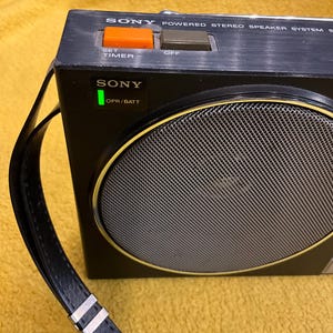 Puede incluir: Un sistema de altavoces est&eacute;reo alimentado por Sony negro con una correa de cuero negro, una hebilla de metal plateado y un altavoz redondo con una rejilla plateada. El altavoz tiene una luz verde que dice "OPR/BATT" y dos botones que dicen "SET TIMER" y "OFF".