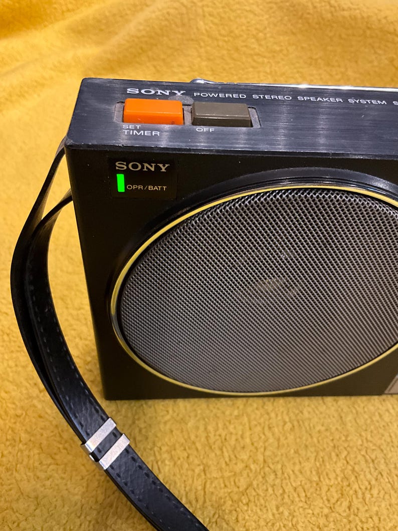 Puede incluir: Un sistema de altavoces est&eacute;reo alimentado por Sony negro con una correa de cuero negro, una hebilla de metal plateado y un altavoz redondo con una rejilla plateada. El altavoz tiene una luz verde que dice "OPR/BATT" y dos botones que dicen "SET TIMER" y "OFF".