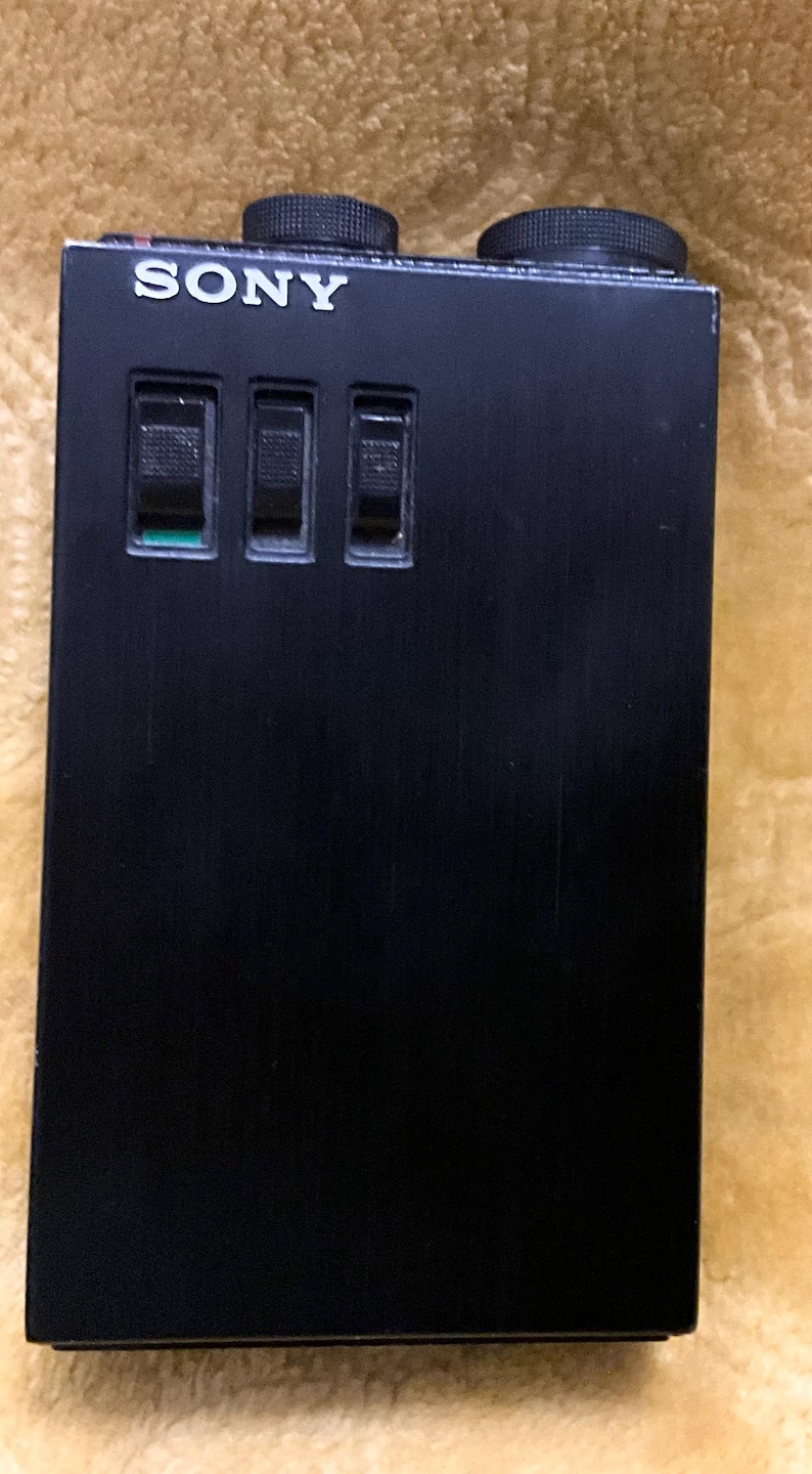 Puede incluir: Un reproductor de casetes Sony negro con tres botones y un logotipo plateado.