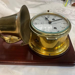 May include: A brass ship clock with a white face and black hands mounted on a rectangular dark wood base. A large brass bell is attached to the side of the clock housing. The clock face features black numerals and a small logo near the center.
