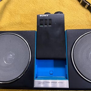 Puede incluir: Un boombox Sony negro con un borde azul y una correa negra. El boombox tiene dos altavoces grandes y un panel de control m&aacute;s peque&ntilde;o con la palabra "SONY" impresa en &eacute;l. El panel de control tiene tres botones y una peque&ntilde;a abertura rectangular. El boombox tiene una etiqueta plateada que dice "POWERED STEREO SPEAKER SYSTEM SRF-60W".