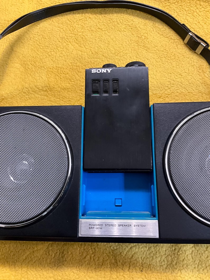 Puede incluir: Un boombox Sony negro con un borde azul y una correa negra. El boombox tiene dos altavoces grandes y un panel de control m&aacute;s peque&ntilde;o con la palabra "SONY" impresa en &eacute;l. El panel de control tiene tres botones y una peque&ntilde;a abertura rectangular. El boombox tiene una etiqueta plateada que dice "POWERED STEREO SPEAKER SYSTEM SRF-60W".