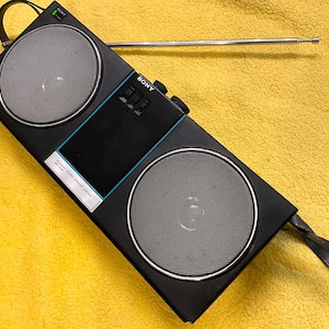 Puede incluir: Una radio est&eacute;reo AM/FM Sony negra con una antena telesc&oacute;pica, dos altavoces y una asa de transporte. La radio tiene una franja azul en la parte delantera y el texto "SONY" y "AM/FM STEREO RADIO" impreso en la parte delantera.