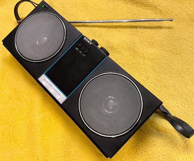 Puede incluir: Una radio est&eacute;reo AM/FM Sony negra con una antena telesc&oacute;pica, dos altavoces y una asa de transporte. La radio tiene una franja azul en la parte delantera y el texto "SONY" y "AM/FM STEREO RADIO" impreso en la parte delantera.