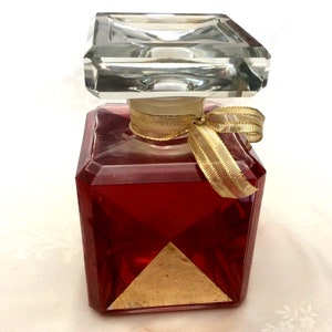 May include: A square glass bottle with a clear glass stopper and a gold ribbon. The bottle is filled with a red liquid and has a gold triangle on the front.