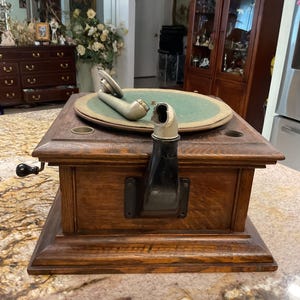 May include: An antique wooden gramophone with a green felt turntable. The gramophone features a metal tone arm and a crank handle. The wooden cabinet has a dark brown finish and sits on a base.