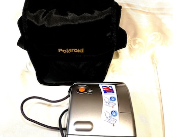 Cámara instantánea Polaroid One plateada con estuche original y película fotográfica en su interior.