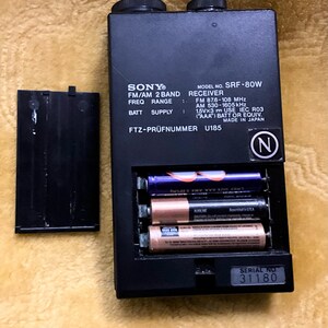 Puede incluir: Un receptor de radio FM/AM de 2 bandas Sony SRF-80W negro con un n&uacute;mero de serie 31180. El receptor tiene un compartimento para bater&iacute;as que contiene tres bater&iacute;as AAA. El receptor tiene una etiqueta en blanco y negro con el texto "FTZ-PR&Uuml;FNUMMER U185" y "MADE IN JAPAN".