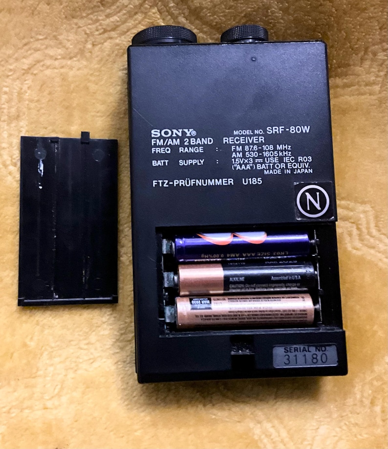 Puede incluir: Un receptor de radio FM/AM de 2 bandas Sony SRF-80W negro con un n&uacute;mero de serie 31180. El receptor tiene un compartimento para bater&iacute;as que contiene tres bater&iacute;as AAA. El receptor tiene una etiqueta en blanco y negro con el texto "FTZ-PR&Uuml;FNUMMER U185" y "MADE IN JAPAN".
