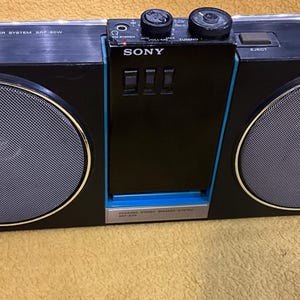 Puede incluir: Un sistema de altavoces est&eacute;reo port&aacute;til Sony negro con un ribete azul. El sistema tiene dos altavoces grandes, un reproductor de casetes y un sintonizador de radio. Tambi&eacute;n tiene una asa de transporte y una correa para el hombro.