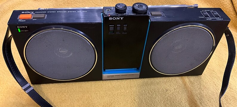 Puede incluir: Un sistema de altavoces est&eacute;reo port&aacute;til Sony negro con un ribete azul. El sistema tiene dos altavoces grandes, un reproductor de casetes y un sintonizador de radio. Tambi&eacute;n tiene una asa de transporte y una correa para el hombro.